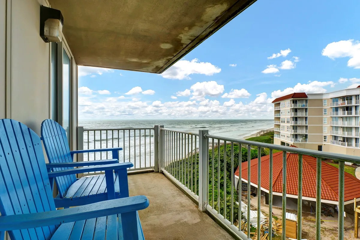 This exterior image presents a balcony with blue Adirondack chairs facing an expansive ocean view under a cloud-filled sky. The scene evokes relaxation, with potential for morning coffee, evening cocktails, or simply unwinding after a productive day.