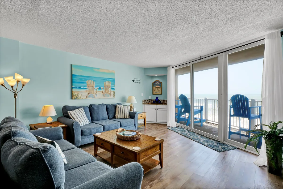 A bright, beach-themed living area with a comfortable sofa, wooden coffee table, and large windows framing an ocean view. The decor is casual and serene, with beach-inspired artwork and ample space for gathering with friends or family.