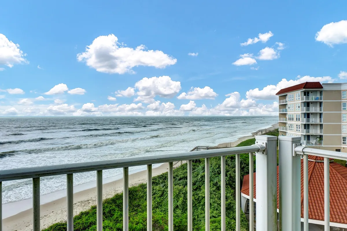 A tranquil scene from the balcony, showcasing the railing, sweeping ocean waves, sandy beachfront, and distant building. The perspective suggests the experience of sitting or standing on the private balcony, soaking in the oceanfront panorama with room for relaxing or sunset cocktails.