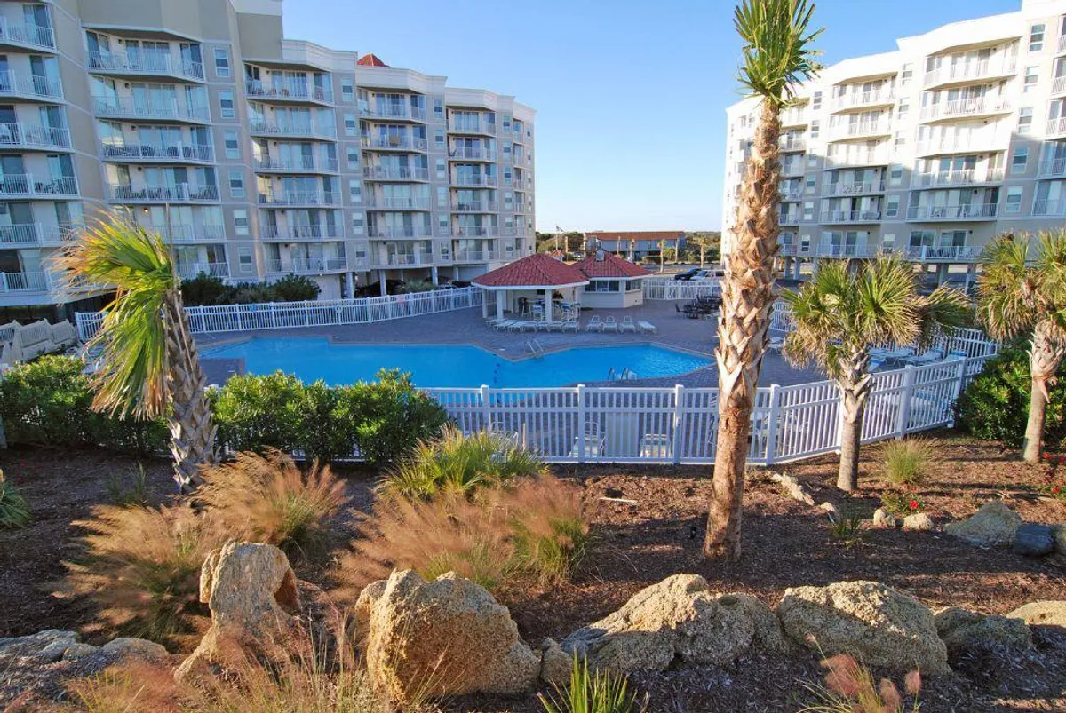This image displays a sparkling swimming pool surrounded by palm trees, with the resort building and gazebo visible in the background. The scene exudes a leisurely, vacation-like atmosphere and highlights sunny weather, poolside relaxation, and spaces for social gatherings.
