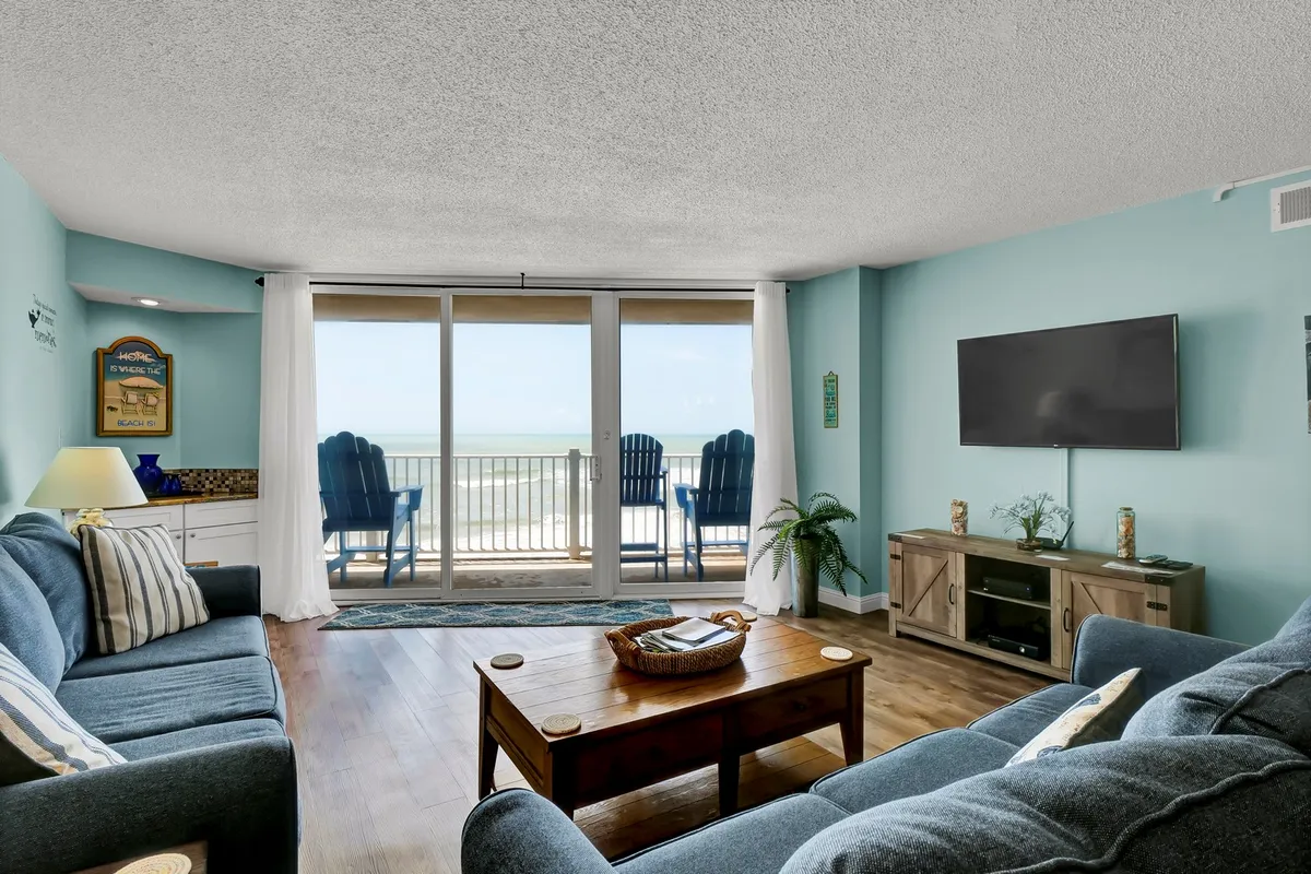 A relaxed living room scene featuring a sofa and coffee table positioned to take in the expansive ocean view through sliding glass doors. The soft natural light highlights the inviting comfort of the space, and the setup suggests it’s ideal for relaxing, socializing, or enjoying entertainment on the TV.
