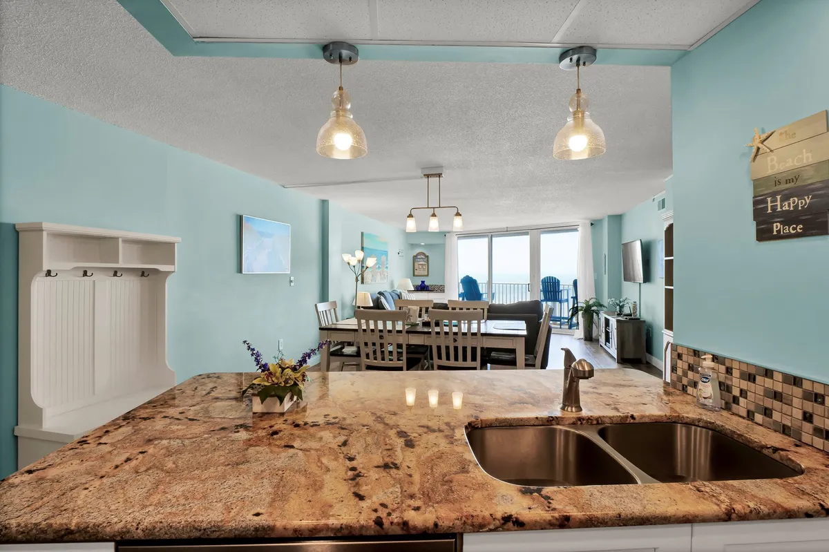 This image showcases a kitchen island with a granite countertop adjacent to a dining area that boasts ocean views through large windows. Decorative touches, such as a wall sign, add a cheerful flair, while the space is oriented for both cooking and entertaining.