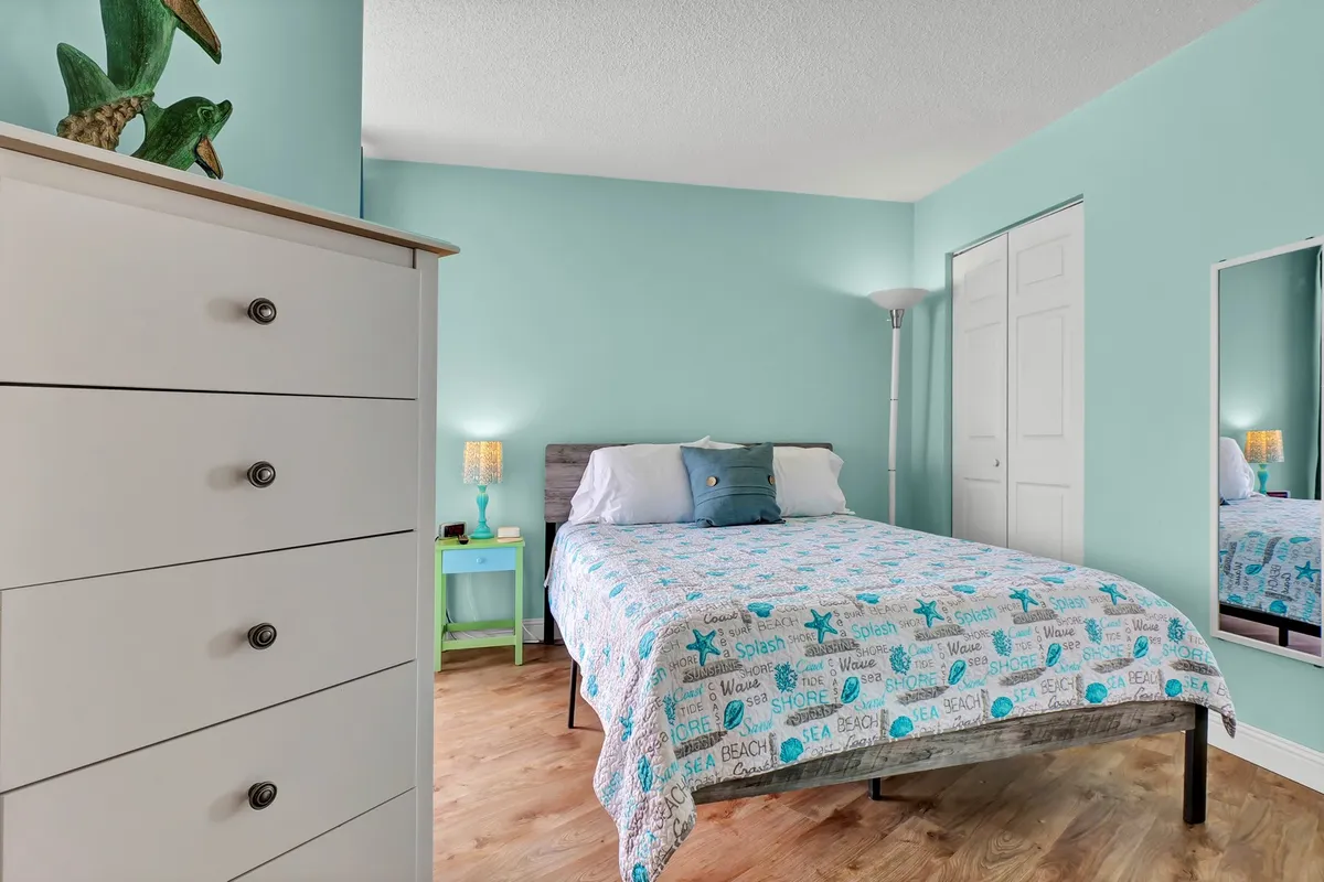 A cozy bedroom with a queen bed dressed in beach-themed bedding, a mirror, nightstand with lamp, and drawer chest. The room feels bright and inviting, suggesting a peaceful place to unwind and recharge.