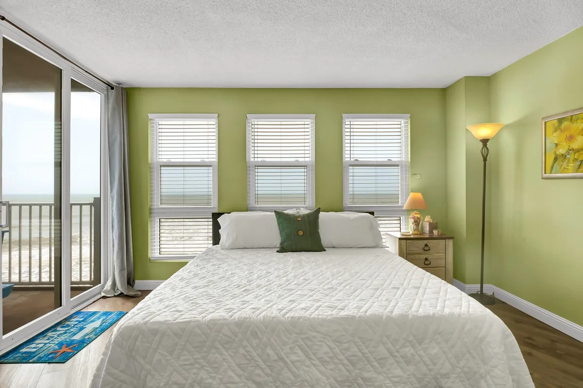 A serene master bedroom featuring a large bed, windows that overlook the ocean, a bedside lamp, and coastal-themed artwork. The space exudes calm and comfort, inviting relaxation and offering scenic views, possibly from a private balcony.