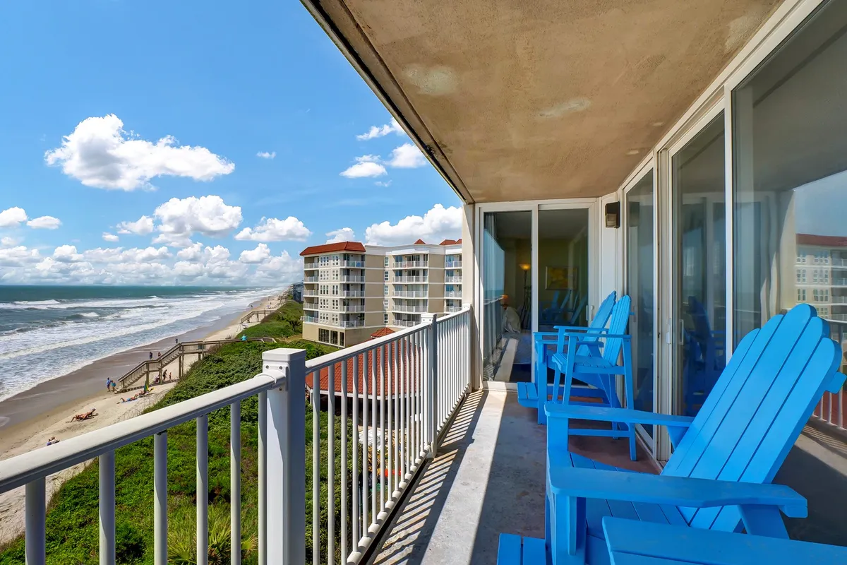 A serene private balcony features two blue Adirondack chairs overlooking the ocean. The scene is tranquil, with soft clouds in the sky and gentle waves, evoking a peaceful atmosphere perfect for unwinding or sharing drinks as the sun sets.