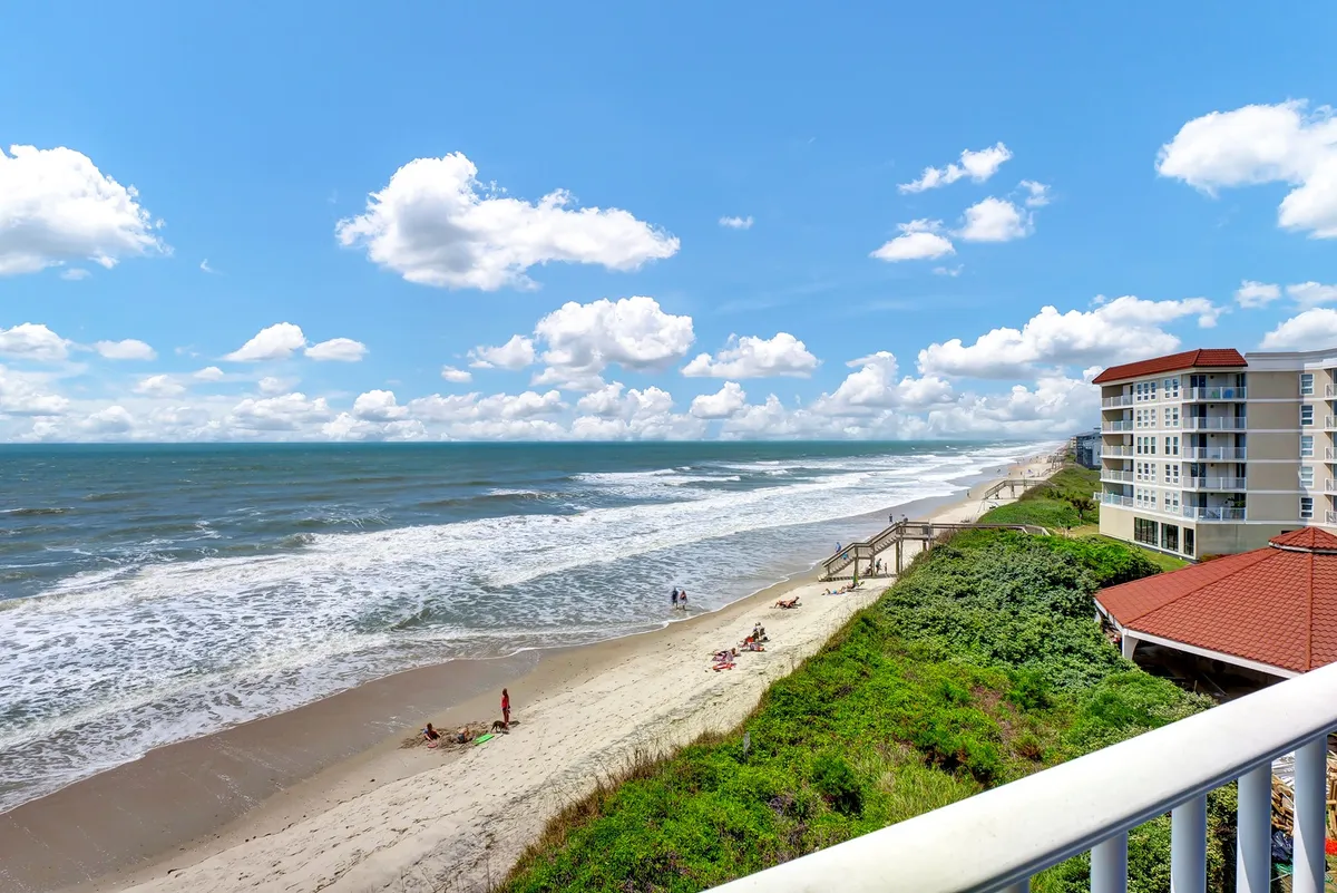 The hero image features sweeping views of the Atlantic Ocean, soft sandy beaches lined with coastal vegetation, and people relaxing, swimming, and enjoying the sun. Nearby buildings suggest convenient access to both the shoreline and local amenities, while the tranquil scene evokes a laid-back, beach vacation vibe.