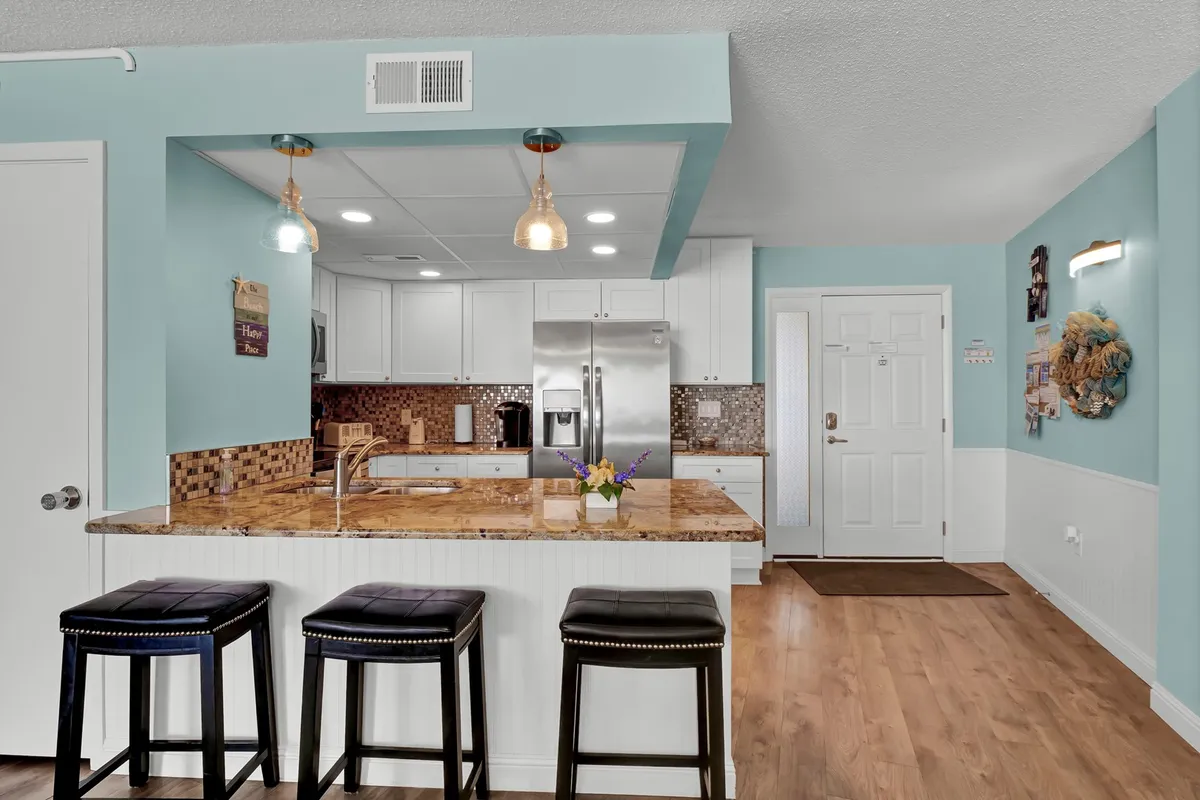 The image displays a welcoming kitchen with granite countertops, bar stools lined along an eat-in counter, full kitchen appliances, and decorative wall art. The space exudes a sense of casual sophistication and is arranged to facilitate both cooking and socializing, with ample space for meal preparation and light conversation.