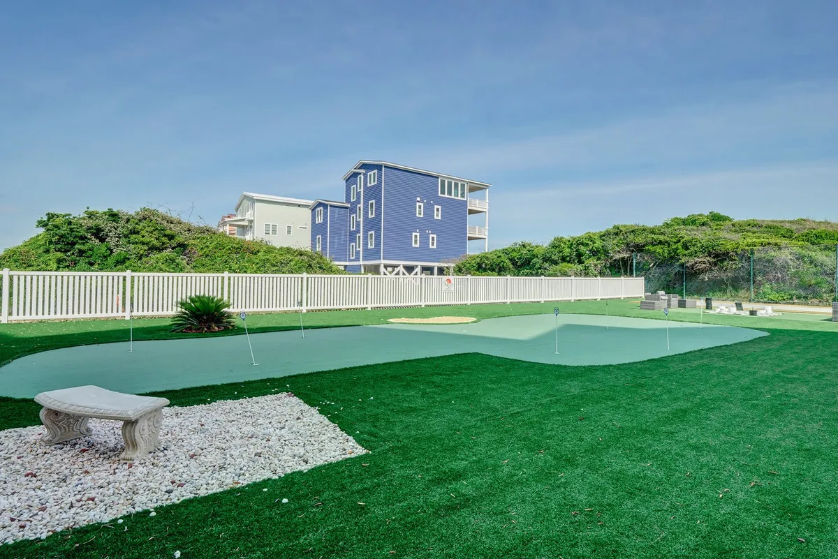 This image highlights a well-manicured putting green bordered by white stones and surrounded by lush greenery and a blue house in the background. The mood is leisurely and social, perfect for family activities or friendly competitions.