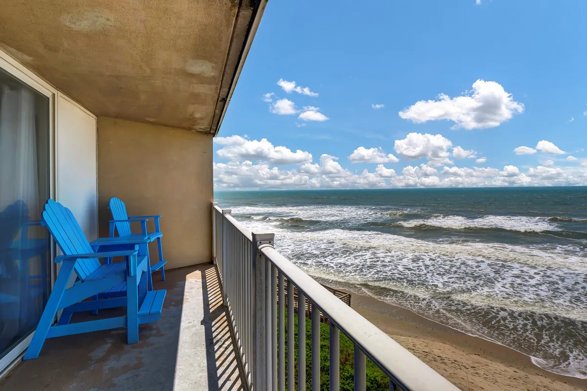 This exterior image features a pair of blue chairs on a balcony overlooking the ocean, with gentle waves and a cloudy sky creating a tranquil, rejuvenating atmosphere. The setup suggests a perfect spot for breaks, casual remote work, or unwinding between meetings while enjoying inspiring sea views.
