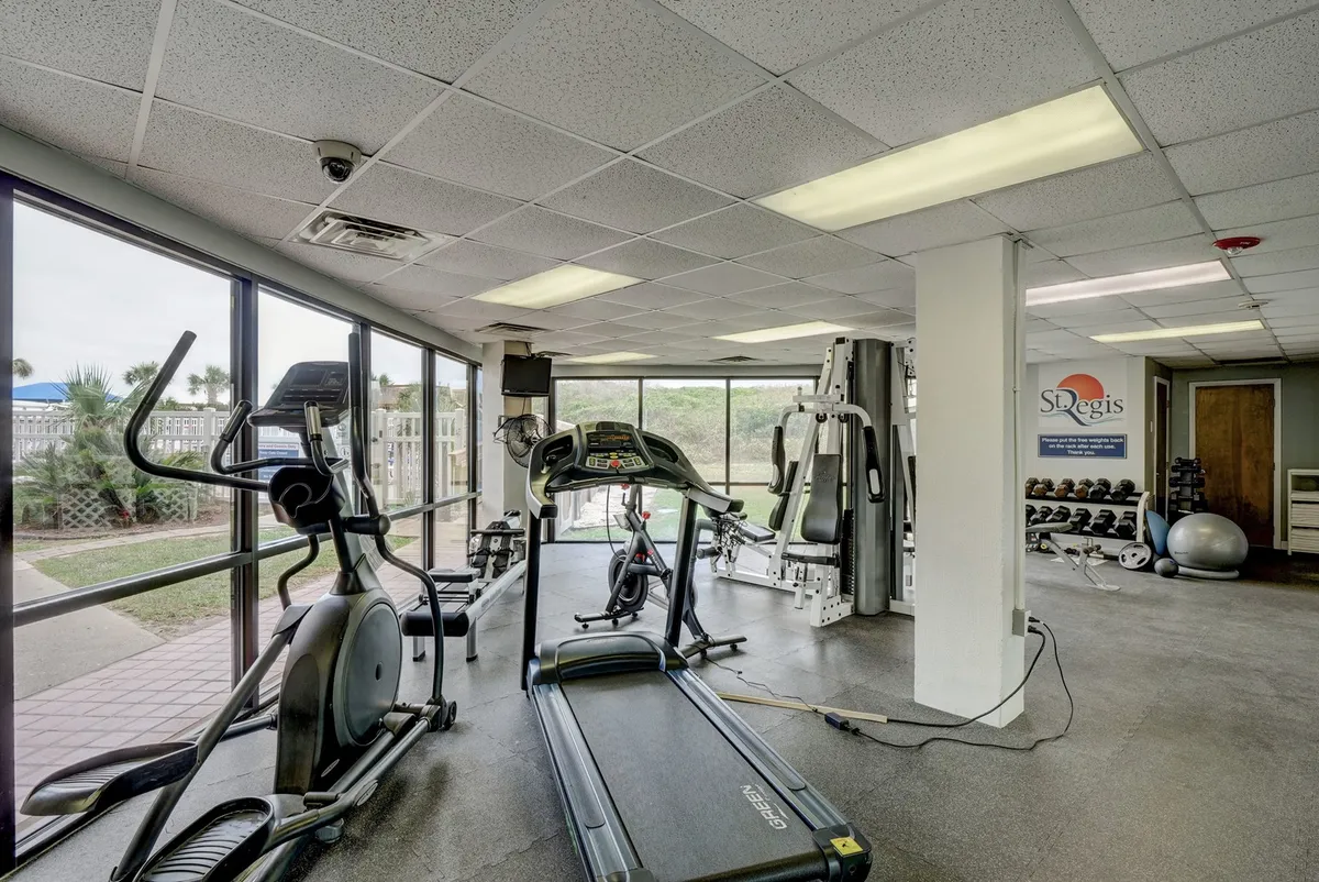 The image features a well-equipped fitness center with a treadmill, elliptical machine, weight training equipment, and large windows allowing natural light to flood the space. The setting is both motivating and inviting, ideal for wellness routines.