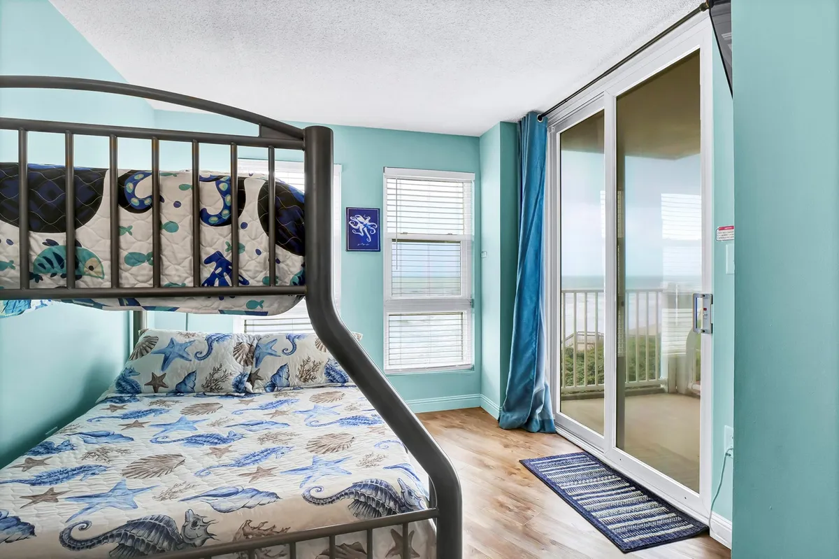A playful bedroom with a twin-over-full bunk bed adorned with colorful bedding, large windows allowing natural light, and an ocean view. The room feels fun and inviting, ideal for children or the young-at-heart.